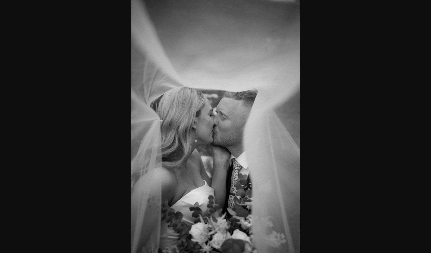 Black and white photograph of a bride and groom kissing under a veil, documentary wedding photography
